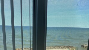 A view through a window with vertical blinds, overlooking a beach with a rocky shoreline and a few scattered chairs.