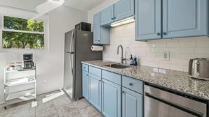 A modern kitchen with light blue cabinets, a stainless steel refrigerator, a granite countertop, and a window with a view of trees. - Belote Place Bungalow (Jacksonville)