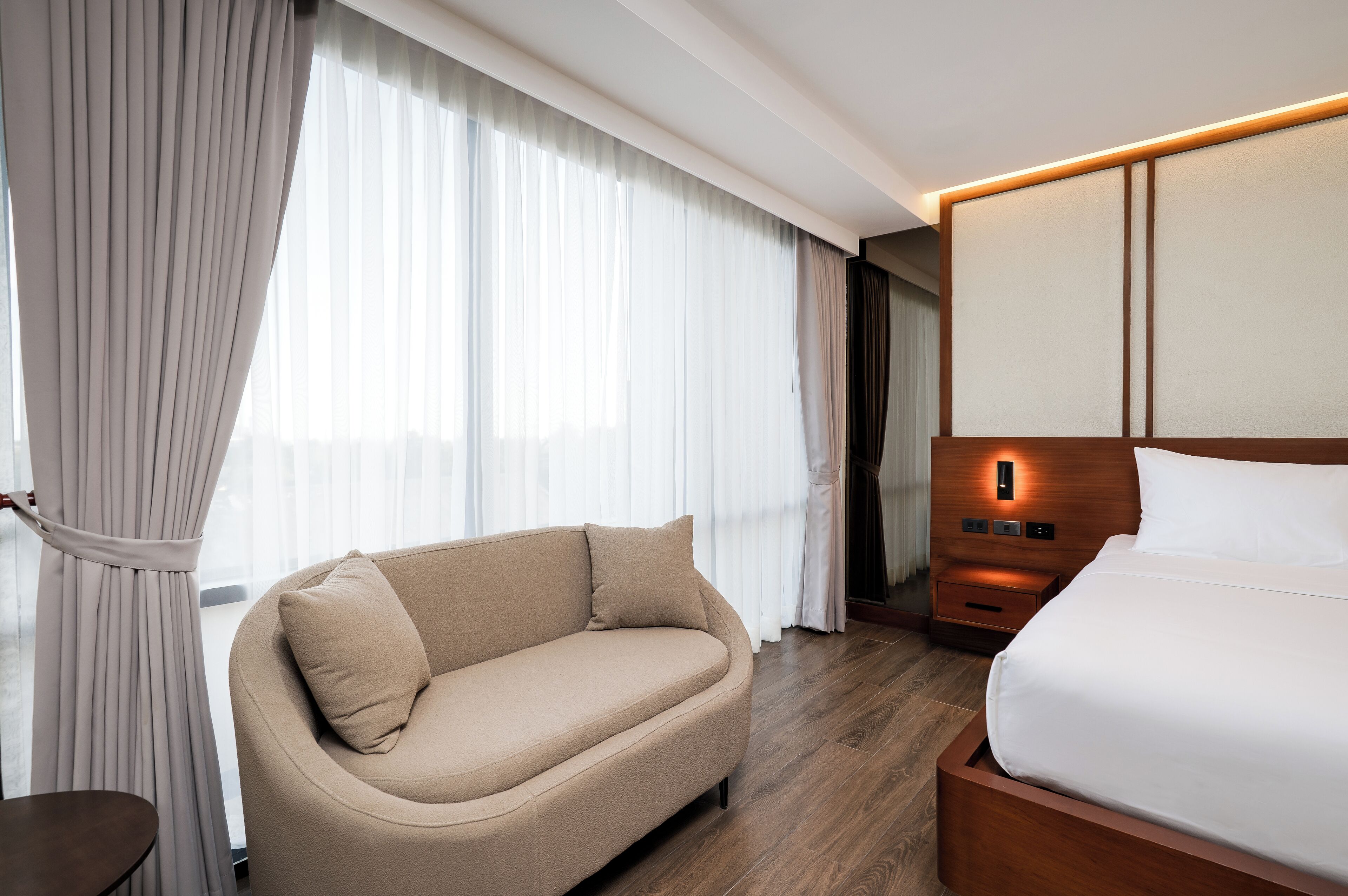 A hotel room with a bed, a sofa, a nightstand, and a large window with curtains.