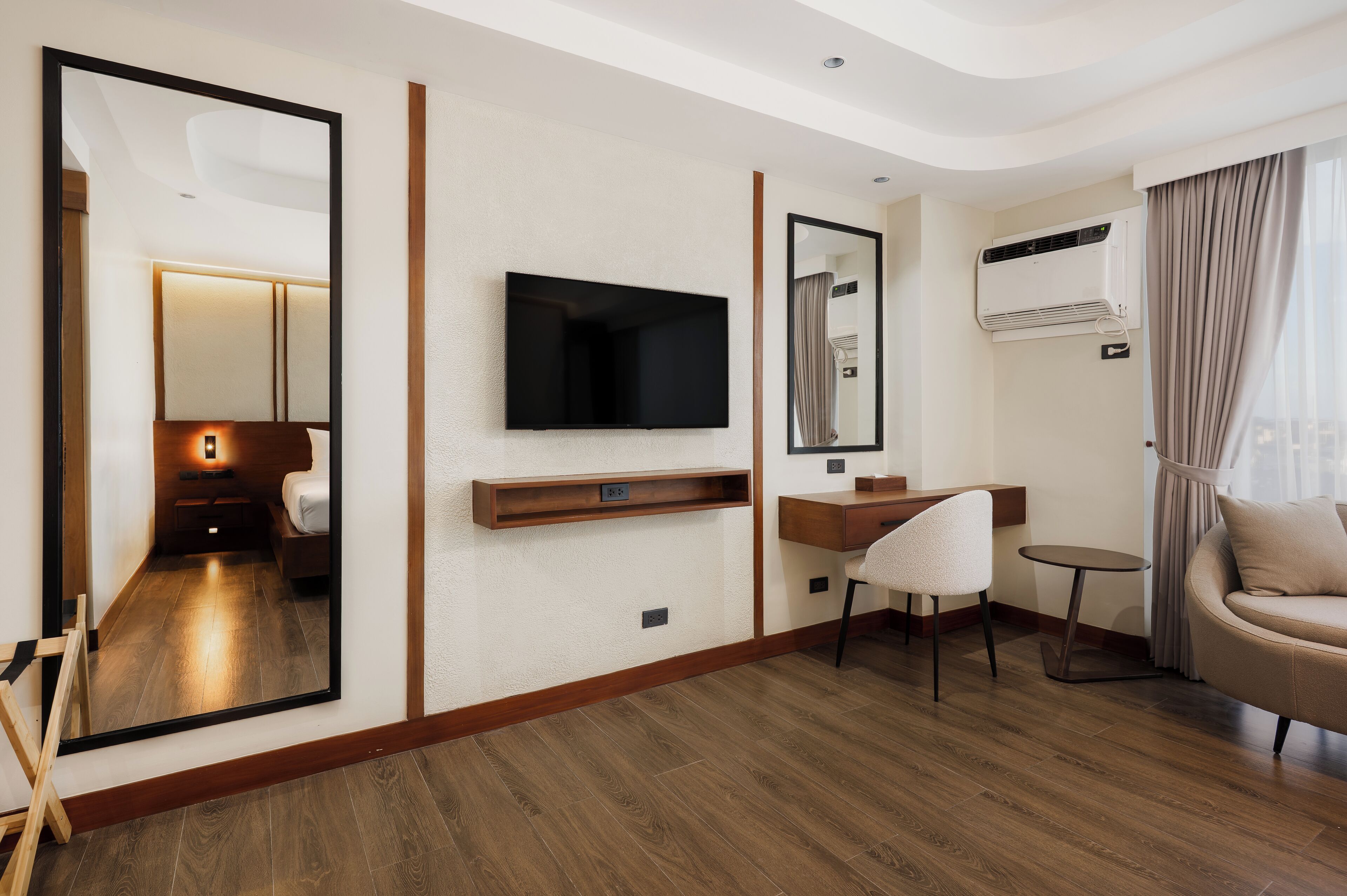 Una habitación de hotel moderna con piso de madera, televisor de pantalla plana, espejo, escritorio, silla, sofá y unidad de aire acondicionado.
