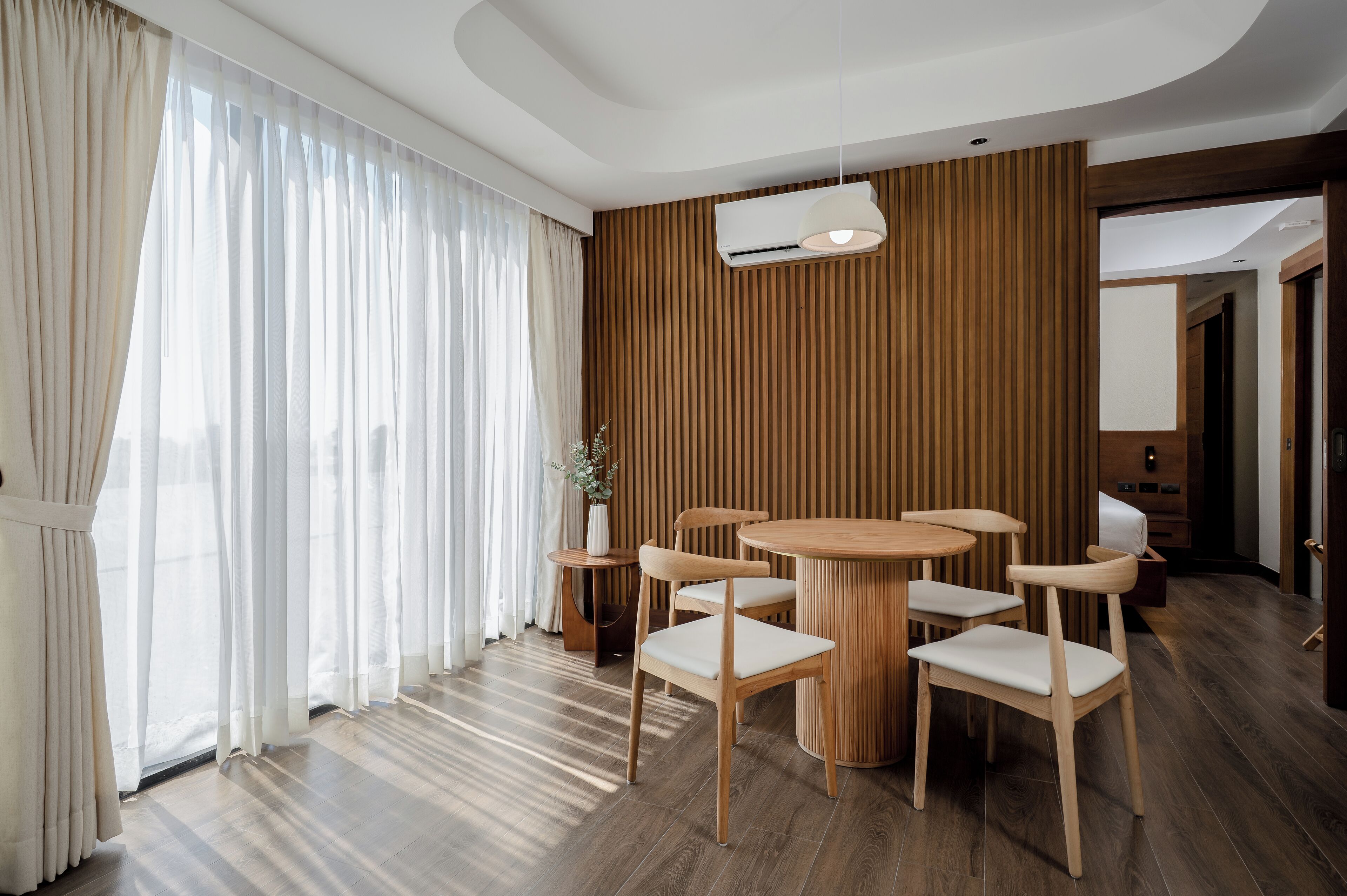 A modern hotel room with a wooden paneled wall, a round wooden table with six chairs, a large window with sheer curtains, and a ceiling-mounted air conditioning unit.
