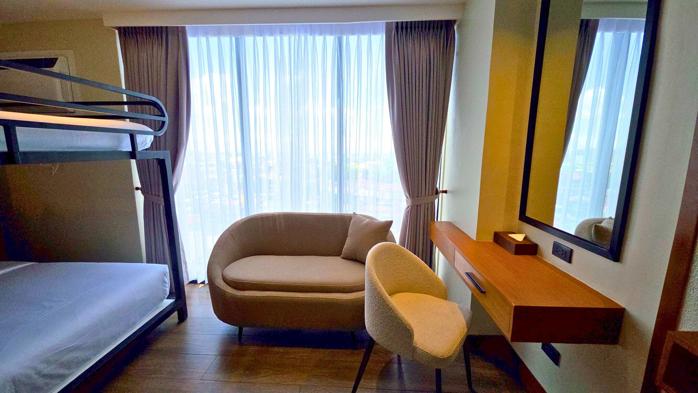 A hotel room with a bunk bed, a sofa, a chair, a desk, a mirror, and a window with curtains.