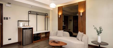 A modern living room with a sofa, coffee table, and a small side table with a plant. There is a kitchenette area with a refrigerator and a wooden cabinet.