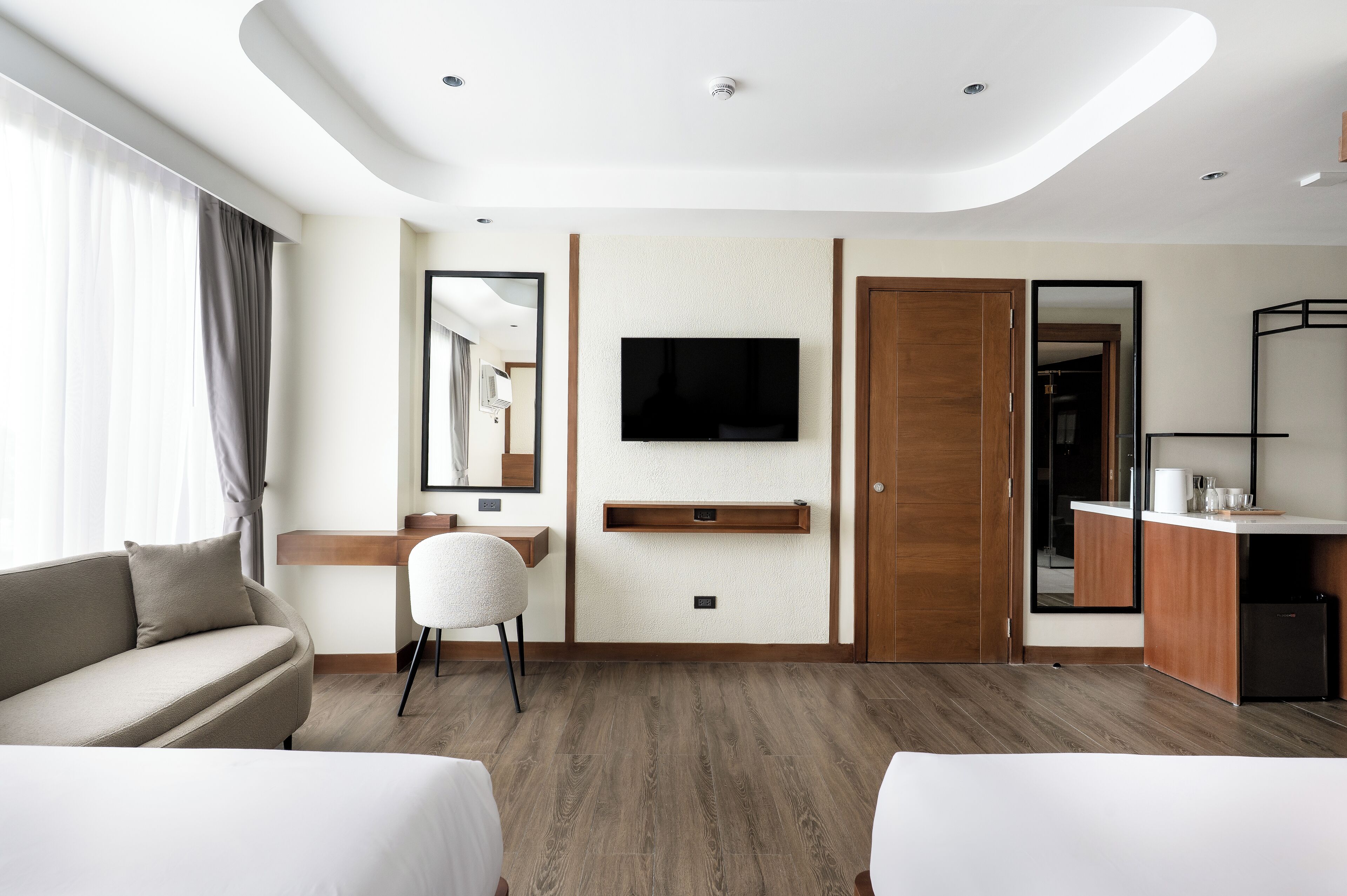 A modern hotel room with a sofa, a chair, a TV mounted on the wall, a wooden cabinet, and a mirror.