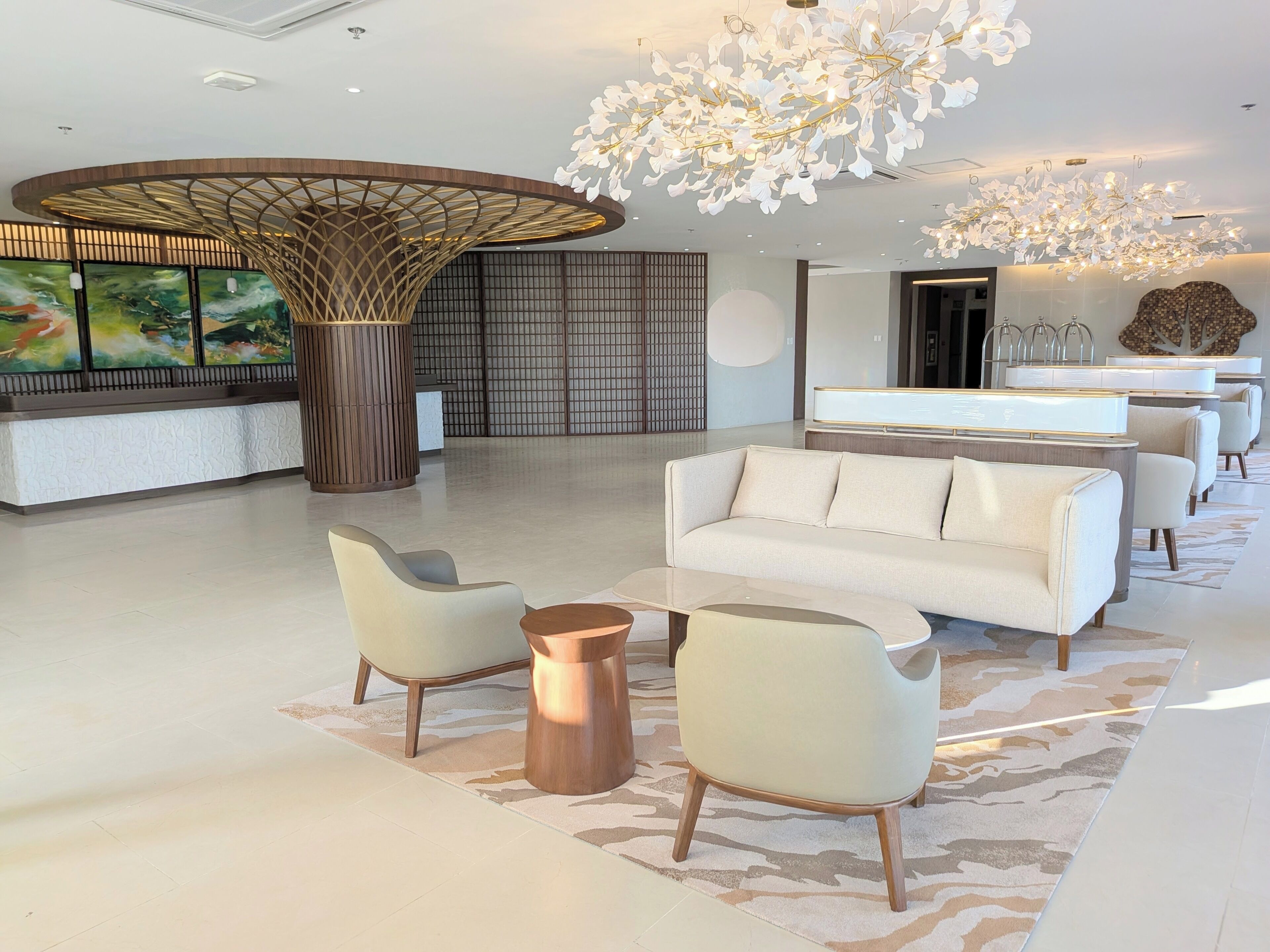 Un moderno vestíbulo de hotel con un sofá blanco, dos sillones, una mesa de centro, una gran lámpara decorativa, un mostrador de recepción y un cuadro de gran tamaño.