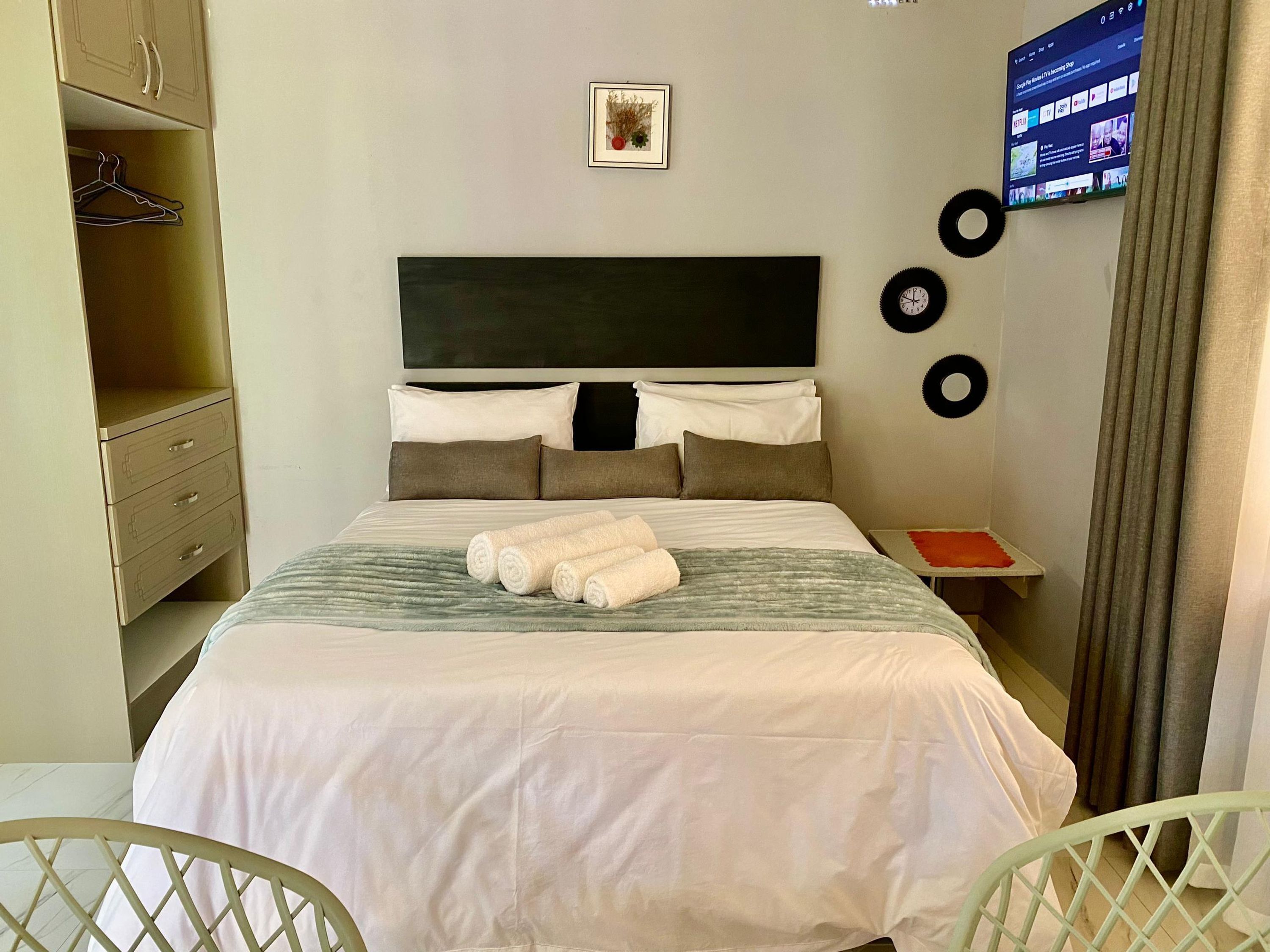 Cama bem arrumada com almofadas e toalhas enroladas, mesa de cabeceira com um objeto vermelho, televisão montada na parede e guarda-roupa com cabides.