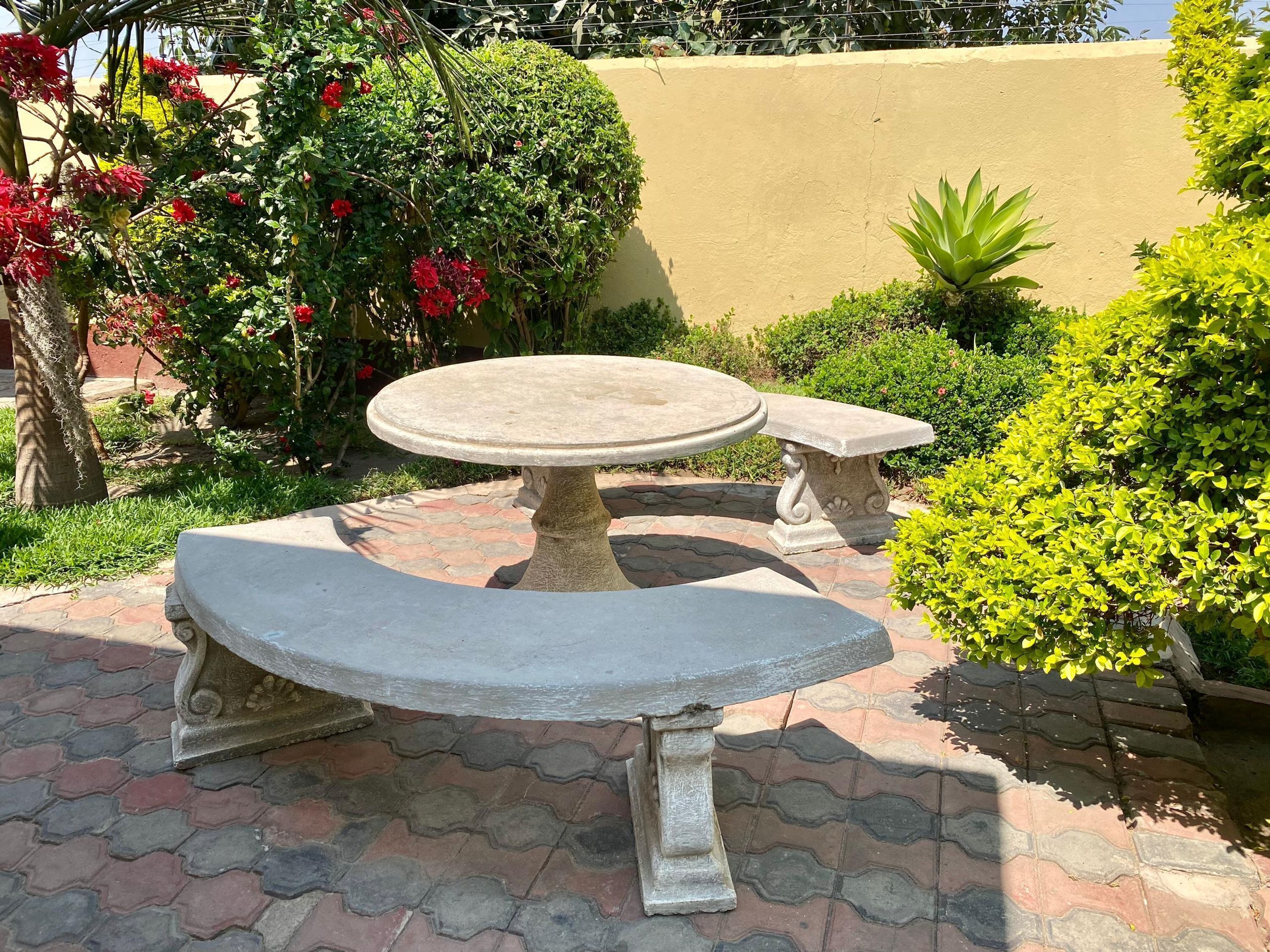 A stone outdoor seating area with a round table and benches, surrounded by lush greenery and a paved walkway.