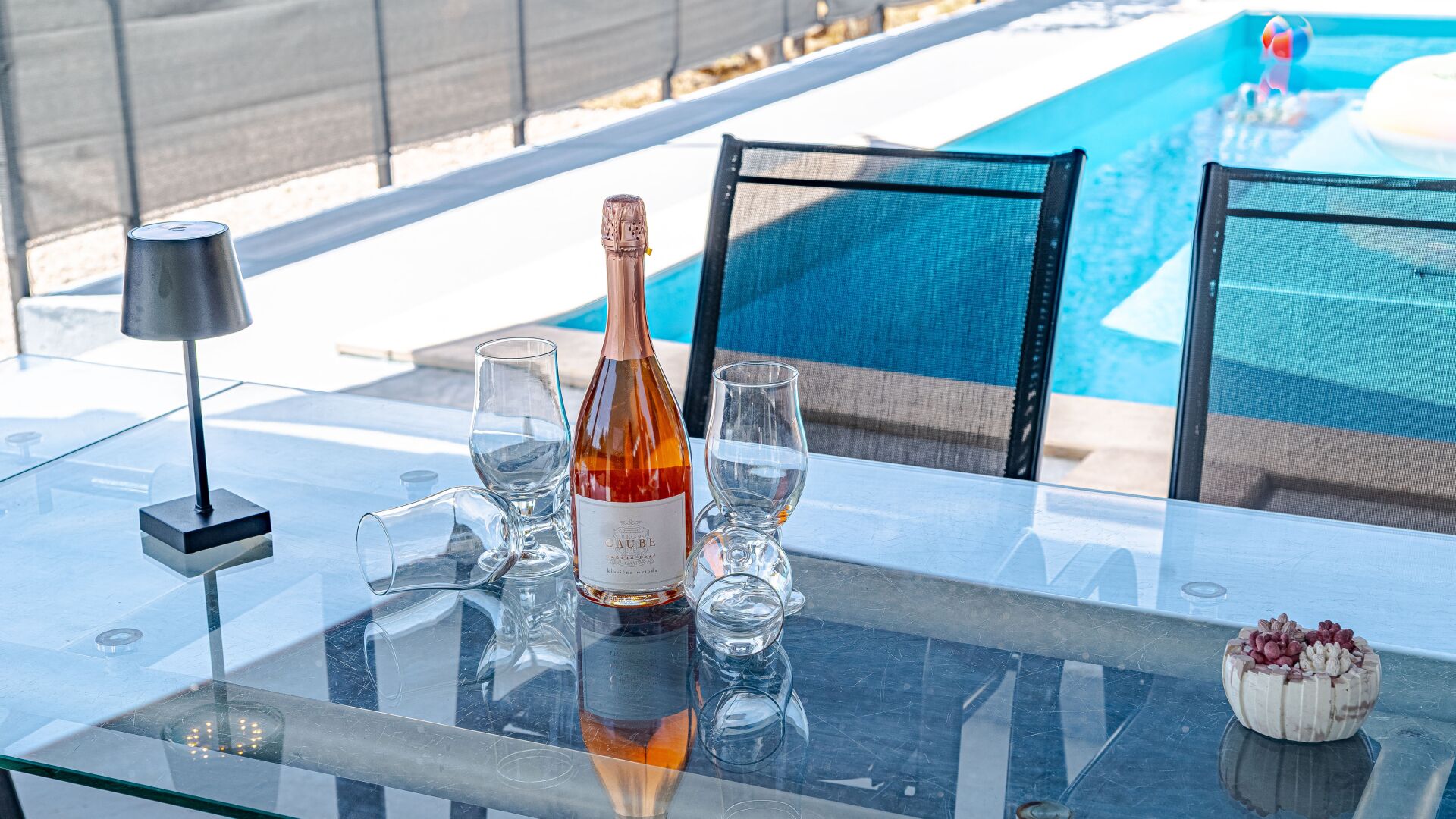 A glass table with a bottle, glasses, and a lamp by a pool.