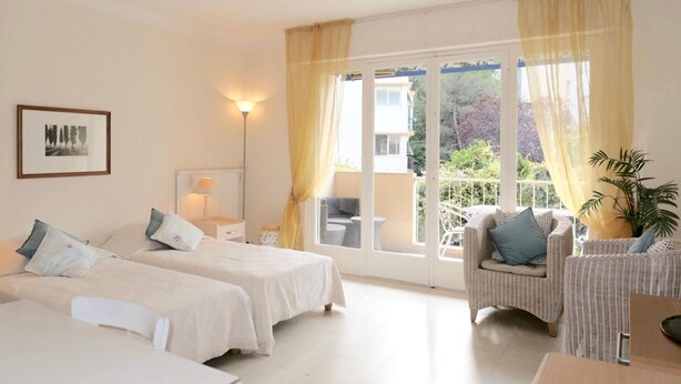 A hotel room with two beds, a sitting area with a chair, a lamp, and a view of greenery outside. - Spacious Studio Apartment Close to The Martinez - 2313 (Cannes)