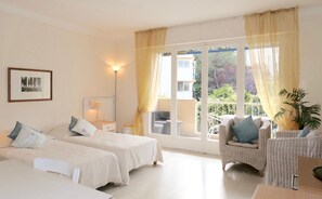 A hotel room with two beds, a sitting area with a chair, a lamp, and a view of greenery outside. - Spacious Studio Apartment Close to The Martinez - 2313 (Cannes)