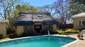A pool area with a thatched-roof structure, a clear pool, and a stone pathway. - Poolside Executive Suite Near Airport - 2322 (Harare)