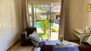 A hotel room with a bed, a desk, a chair, and a view of a swimming pool and outdoor area. - Poolside Executive Suite Near Airport - 2322 (Harare)