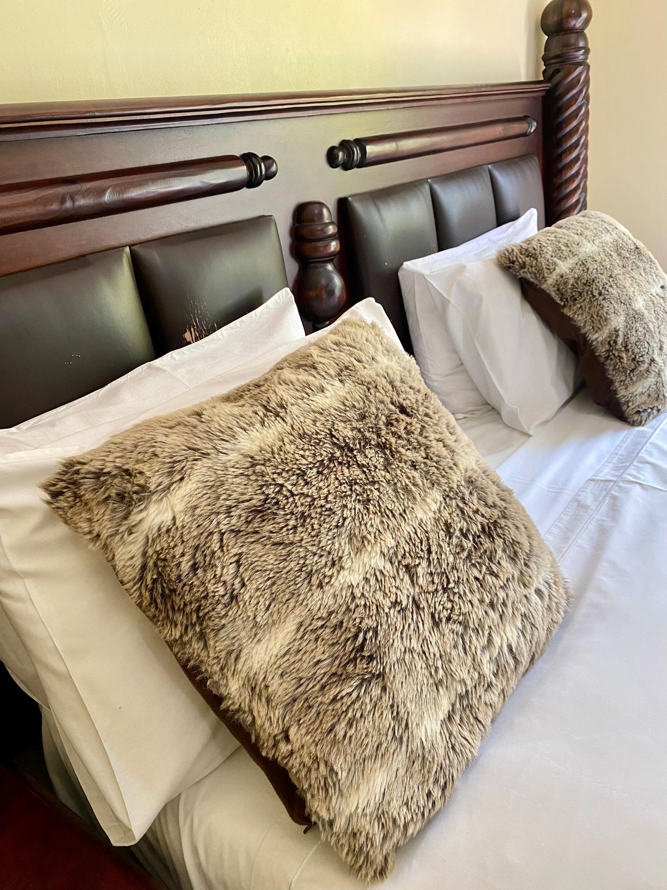 A bed with a dark wooden headboard, white linens, and a large fur throw pillow.