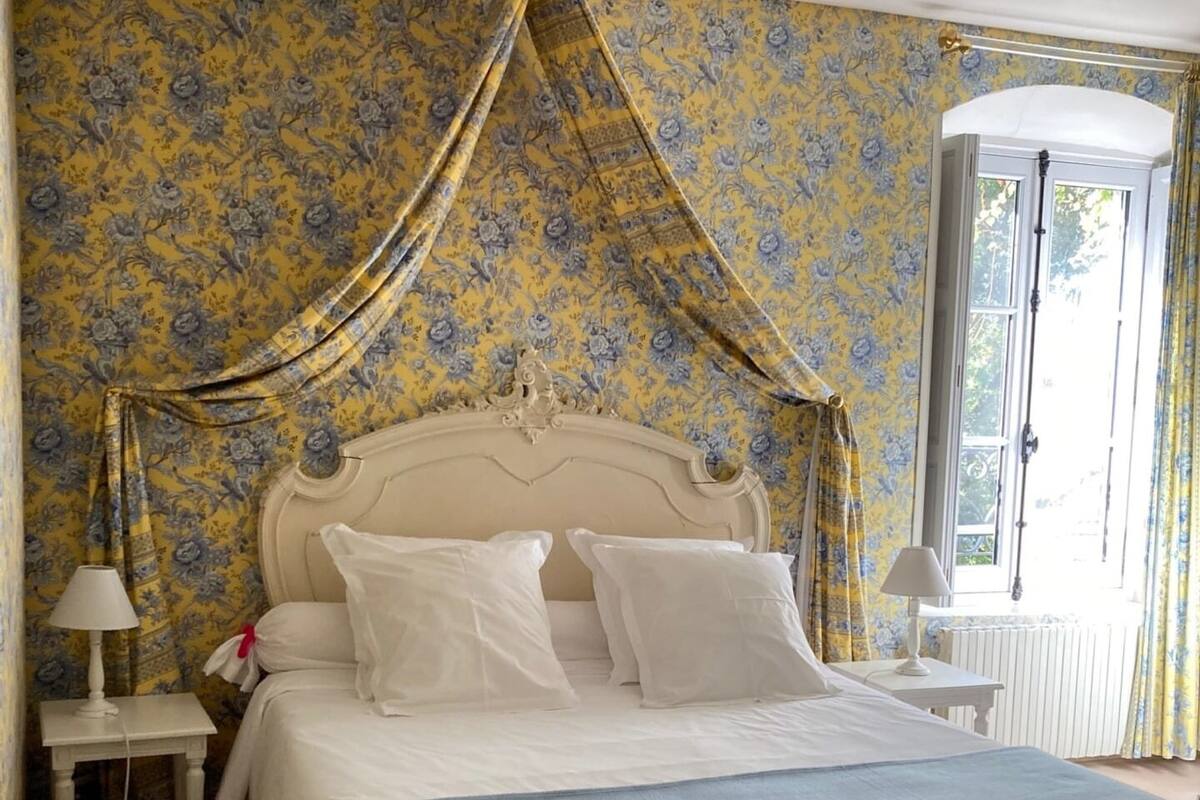 Une chambre à coucher avec un lit à baldaquin, un papier peint à motifs floraux et une fenêtre avec des rideaux.