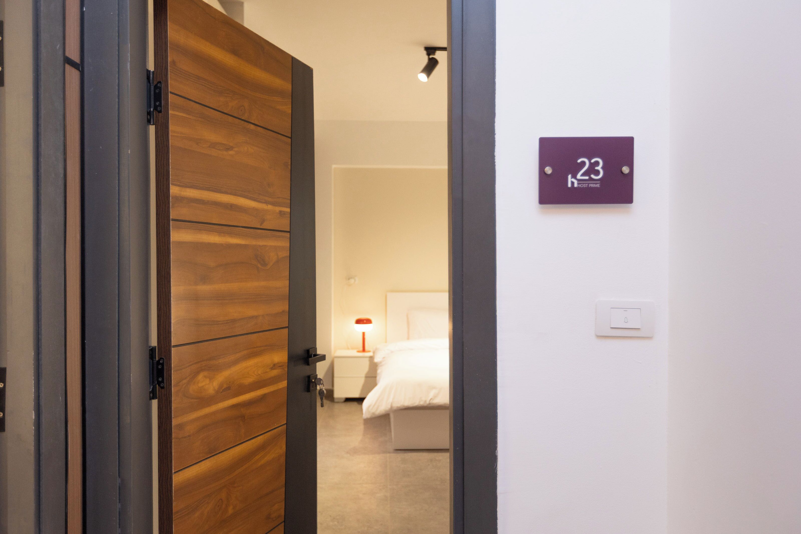 A hotel room with a wooden door, a bed with white bedding, a nightstand with a red lamp, and a wall-mounted sign indicating room number 23.