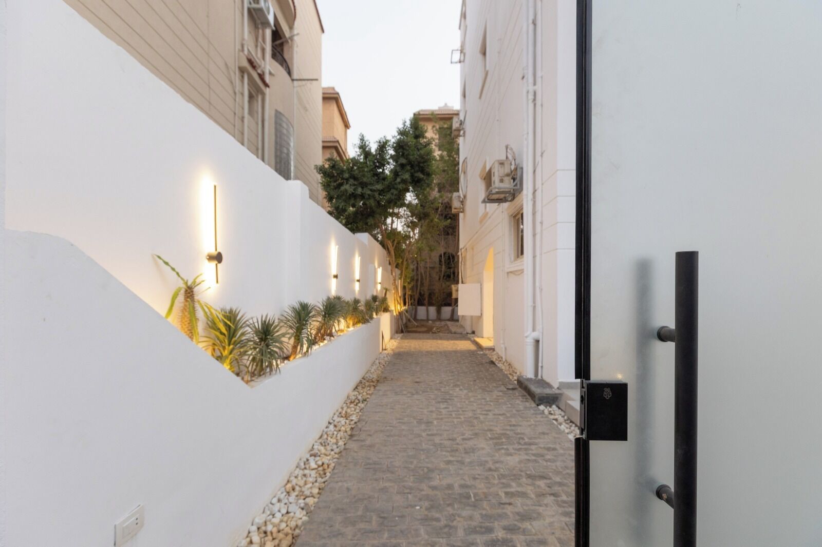 Une étroite ruelle pavée, bordée de murs blancs, agrémentée de luminaires modernes et d’un petit jardinier avec des plantes.