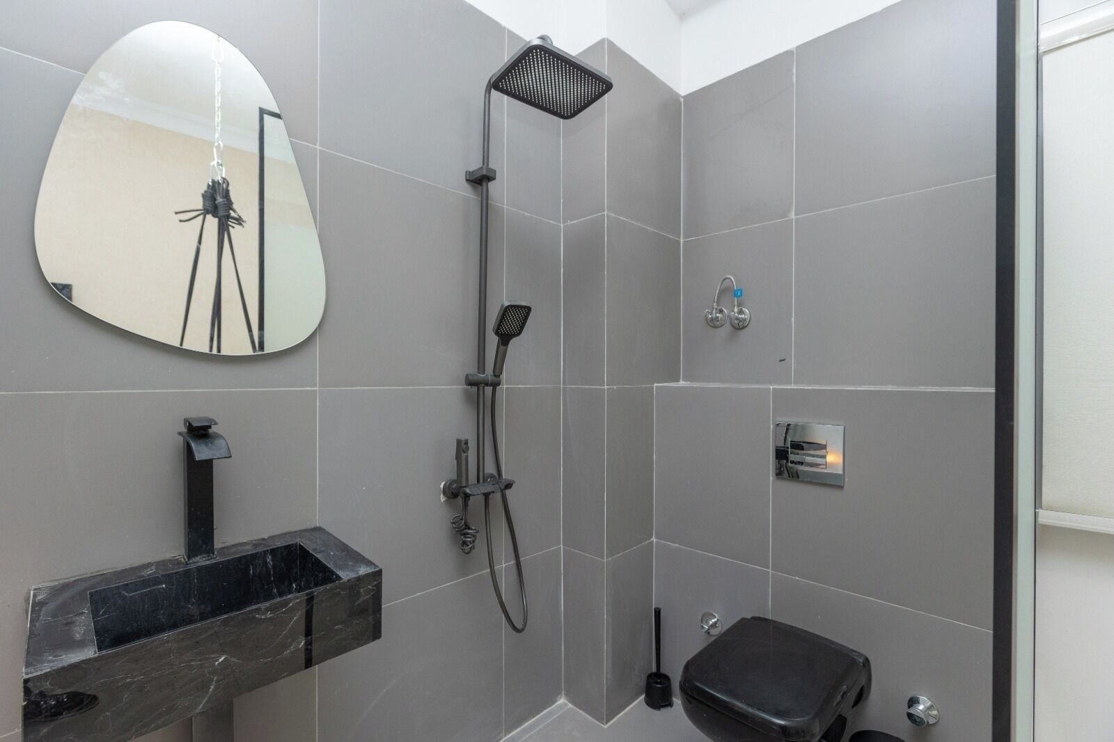 Un baño moderno con azulejos grises, un espejo redondo, un lavamanos de mármol negro, un cabezal de ducha manual y un inodoro.