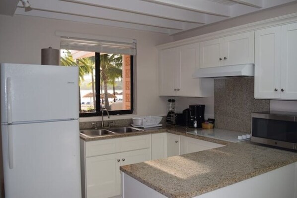 A kitchen with white cabinets, a granite countertop, a refrigerator, a sink, and a window with a view of palm trees. - Beautiful Beach Front Villa in a prime location (Saint Paul's)
