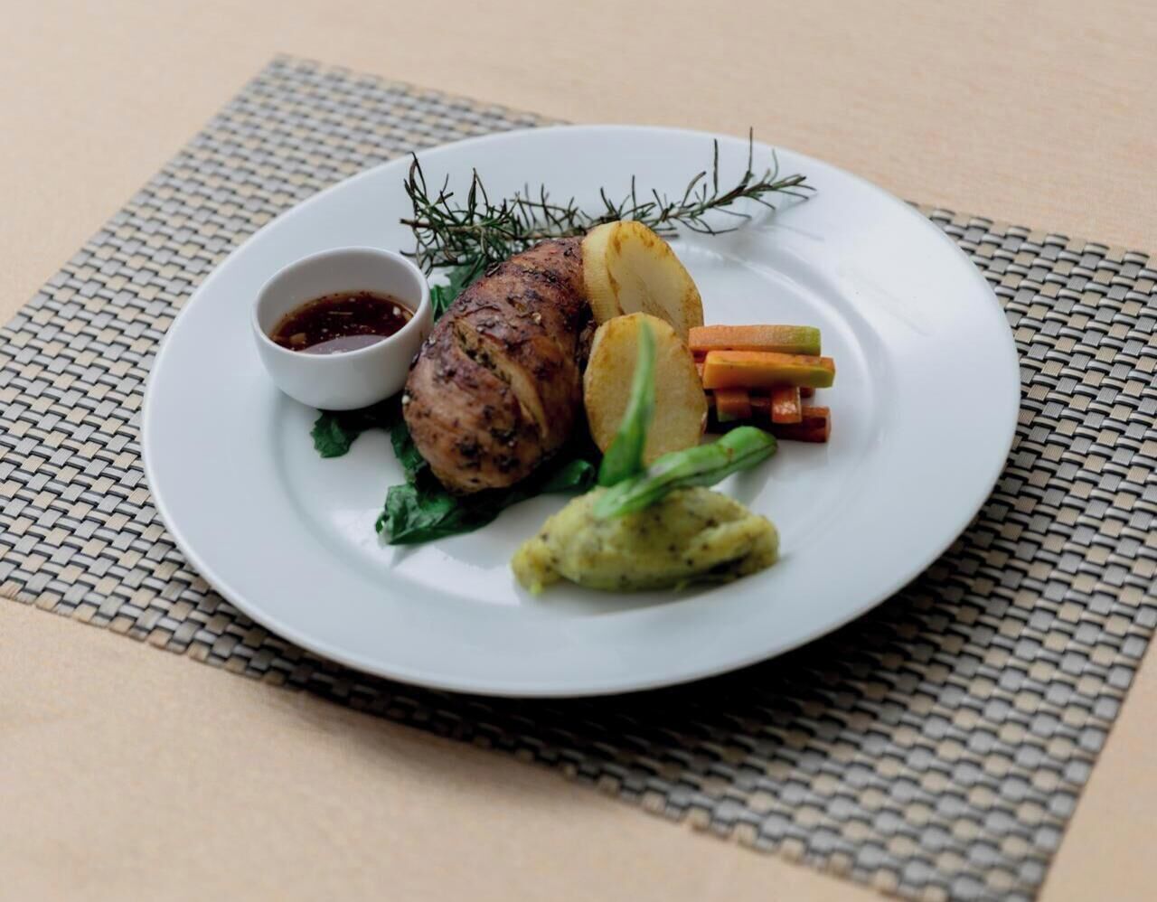 A plate with a roasted meat, vegetables, and a small bowl of sauce on a woven placemat.