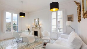 Amplia sala de estar con un sofá blanco, una mesa de vidrio y dos sillas blancas. Hay una chimenea con un espejo encima y un balcón con barandilla.