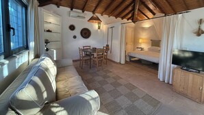 A cozy room with a wooden ceiling, a sofa, a dining table with chairs, a bed with a canopy, and a TV on a wooden cabinet.
