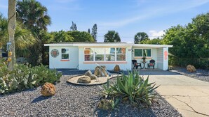 Ein kleines, eingeschossiges Gebäude mit weißer Fassade und grossen Fenstern. Es hat eine Terrassenfläche mit Tisch und Stühlen, umgeben von verschiedenen Pflanzen und Steinen.