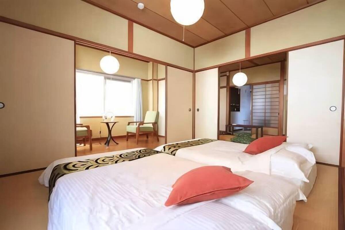 A traditional Japanese-style room with tatami flooring, a low wooden ceiling, and sliding doors. There are two beds with white bedding and red pillows, a small table with chairs, and a view of a traditional Japanese room through the sliding doors.