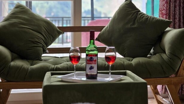 A cozy seating area with two cushioned chairs, a small table with a bottle and glasses of red wine, and a window with a view of greenery. - Hotel New Snow Palace  (Dharamshala)