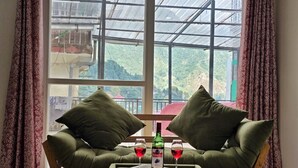 A room with a large window, a green sofa with two pillows, a small table with a bottle and two glasses of red wine. - Hotel New Snow Palace  (Dharamshala)