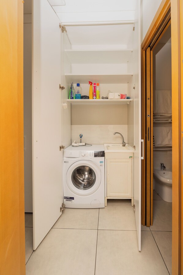 A compact laundry room with a washing machine, a sink, and shelves with cleaning supplies. - La Casa del Nostromo (Salerno)