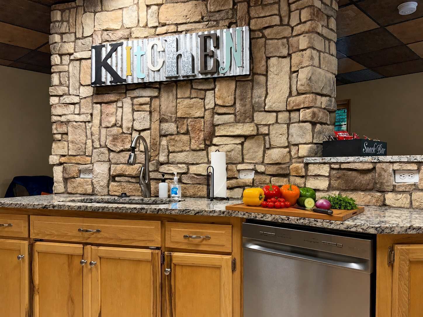 A kitchen with a stone wall, a stainless steel dishwasher, wooden cabinets, a faucet, and a snack bar sign.