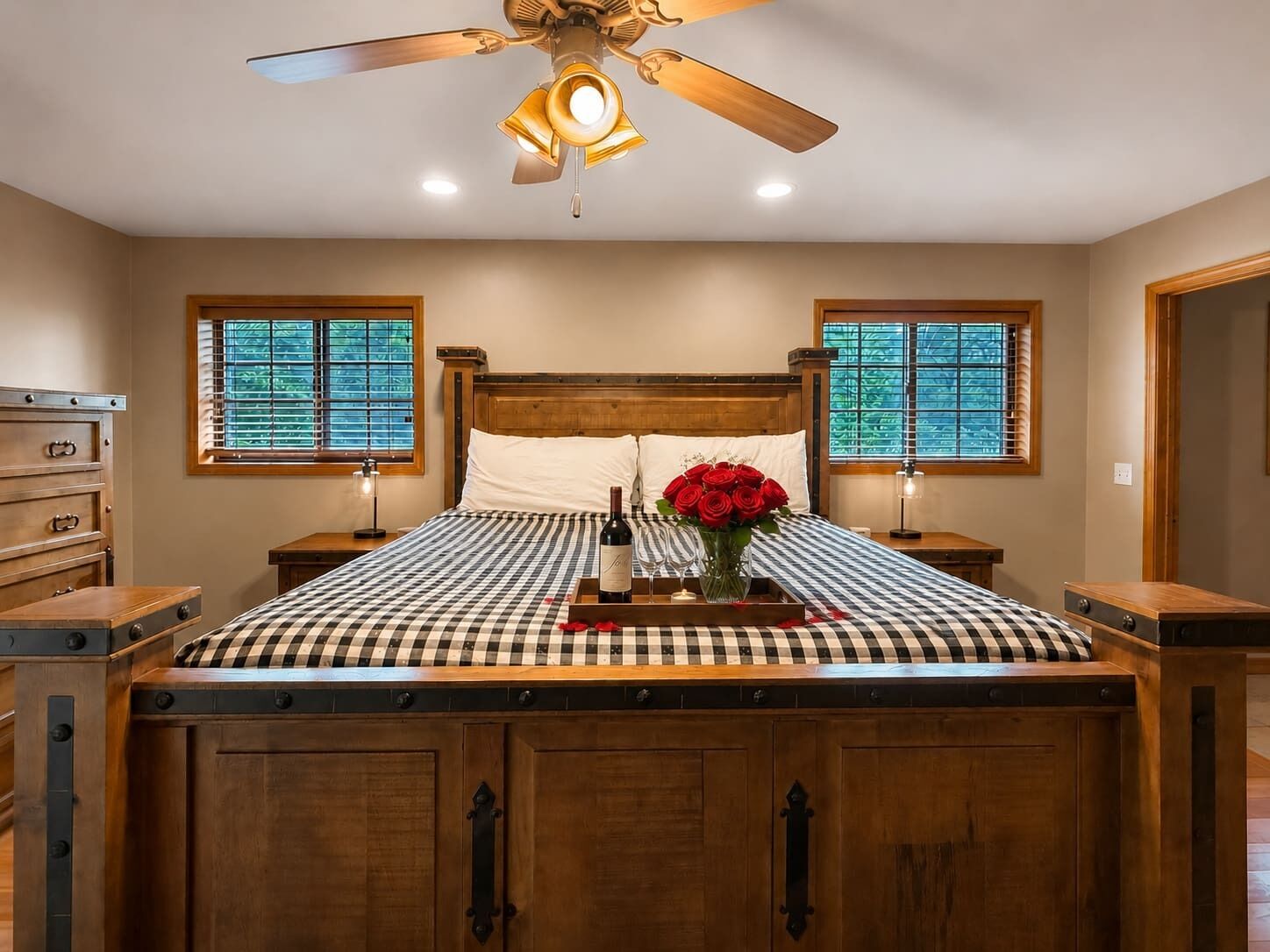 A bedroom with a wooden bed, checkered bedding, a ceiling fan, and two windows with blinds.