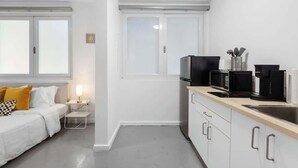 Un espace de vie compact comprenant un coin cuisine, un lit avec une literie blanche et un oreiller jaune, une petite table de chevet avec une lampe, et une fenêtre avec des panneaux de verre dépoli.