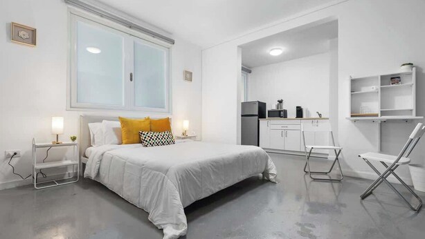 Une chambre moderne avec un grand lit, des tables de chevet, un petit bureau et, au fond, un coin cuisine.