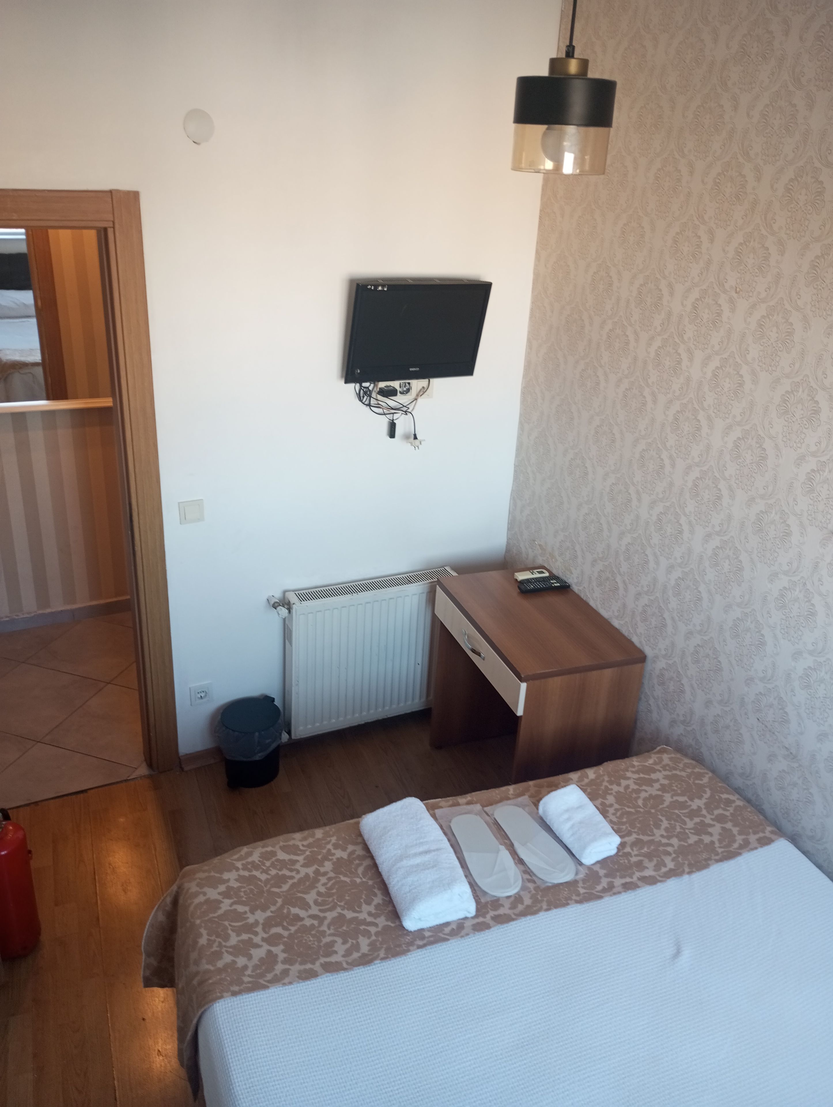 Una habitación de hotel con una cama, un escritorio, un televisor montado en la pared, un radiador y un cubo de basura.