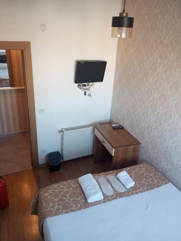 A hotel room with a bed, a desk, a television mounted on the wall, a radiator, and a trash bin. - Kalamaripansiyon2 (Adalar)
