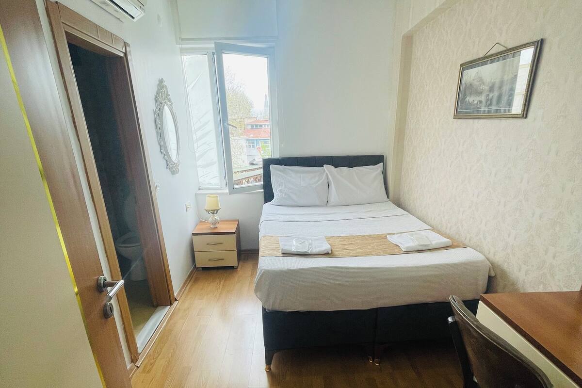 A bedroom with a bed, a desk, a chair, a bedside table, an air conditioner, a window, and a door.