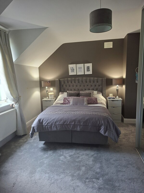Une chambre à coucher avec un grand lit, deux tables de chevet, un luminaire au plafond et une fenêtre avec des rideaux.