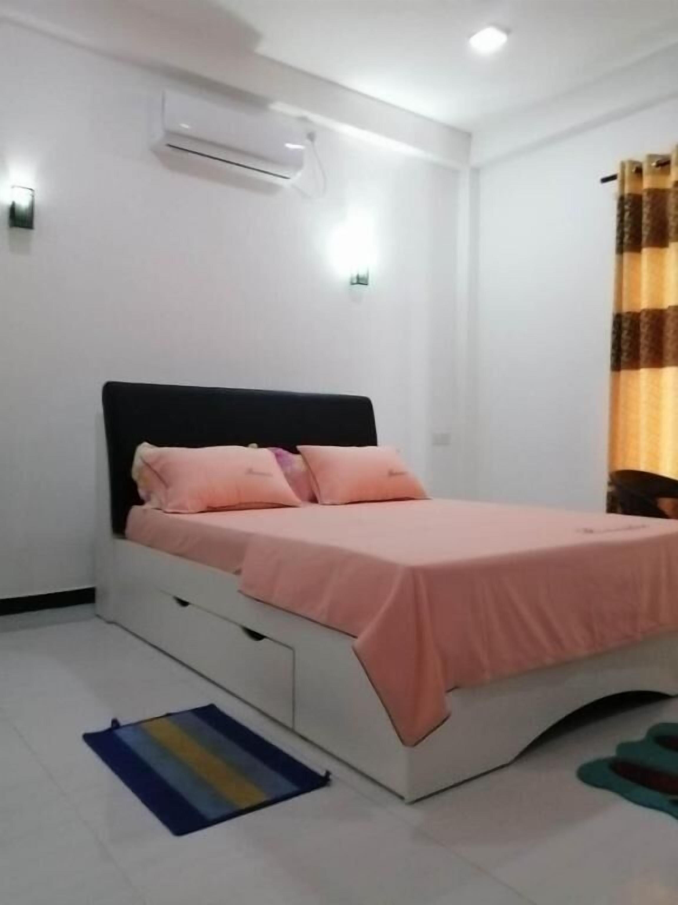 A bedroom with a bed, white walls, an air conditioning unit, and a striped rug.