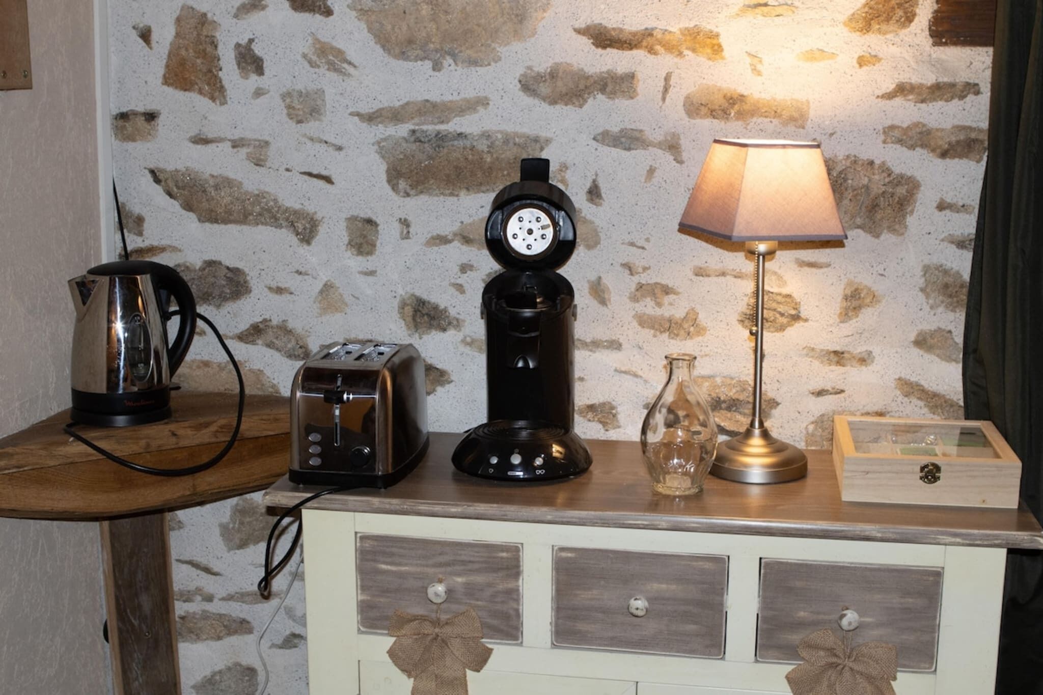 Ett litet kök med en kaffebryggare, brödrost och en lampa placerade på ett träbord framför en stenmur.
