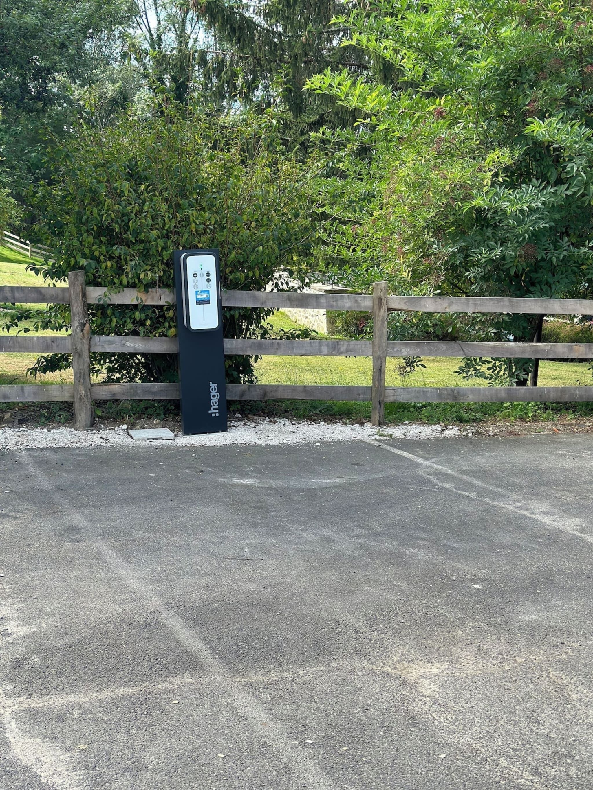 Un espace de stationnement équipé d’une borne de recharge portant l’inscription « Hager », situé près d’une clôture en bois et d’une zone verdoyante.
