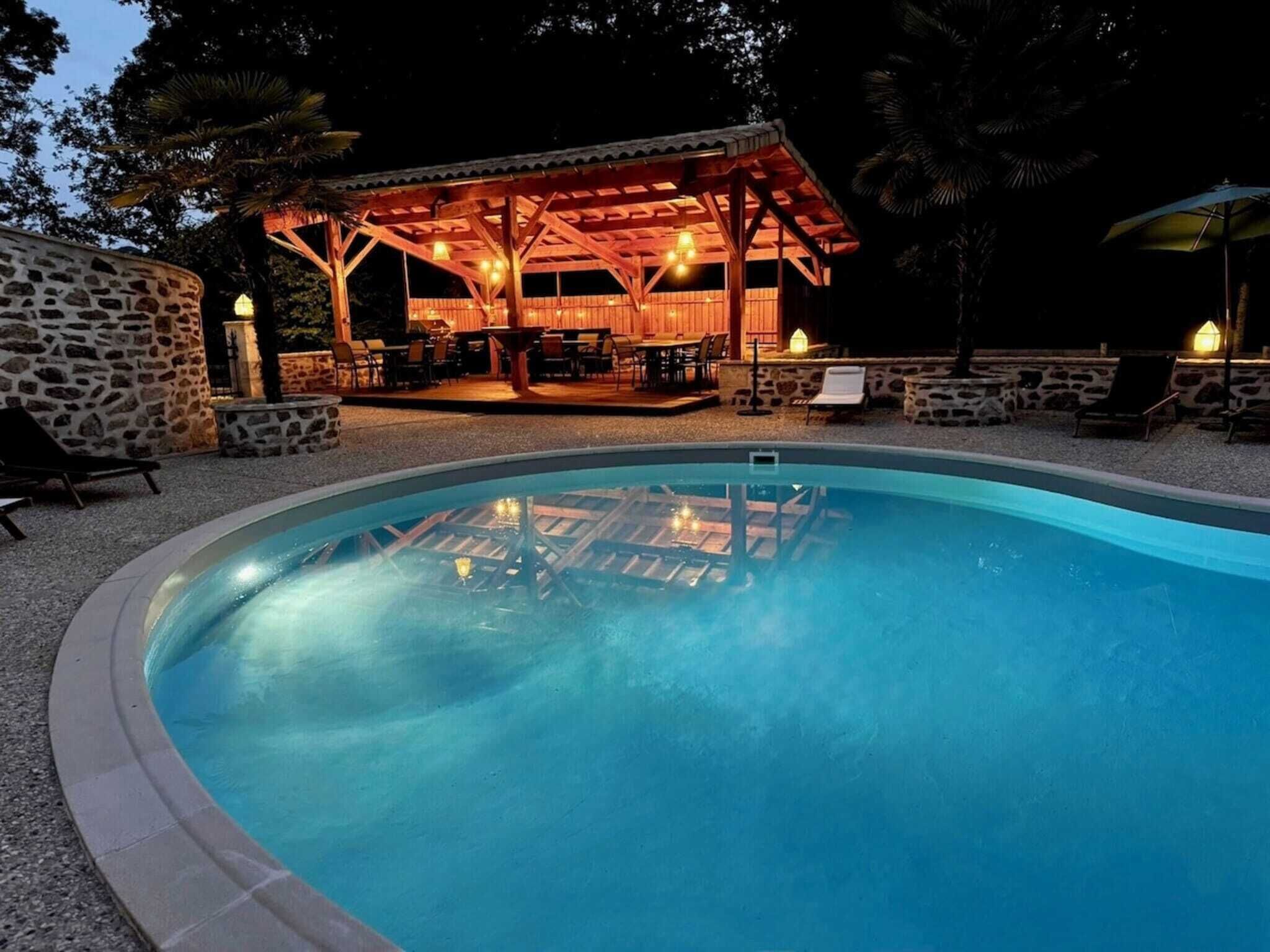 A swimming pool with a wooden deck and lounge chairs, surrounded by stone walls and a covered seating area with tables and chairs.