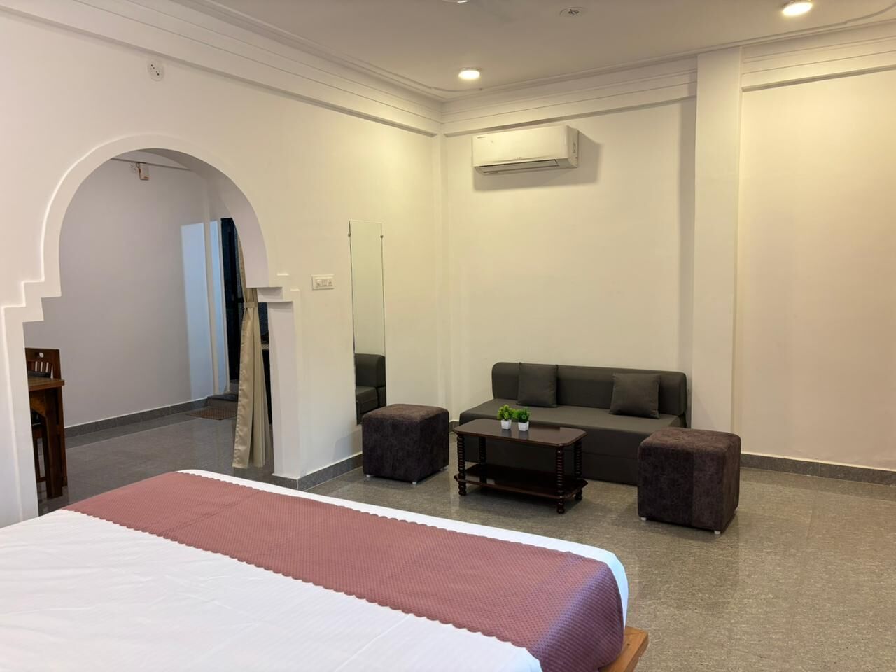 Una camera d'hotel con un letto, un divano, due poggiapiedi, un tavolino con piante e un condizionatore d'aria.