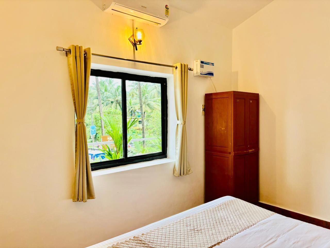 A room with a bed, a window with curtains, a wooden wardrobe, and an air conditioning unit.