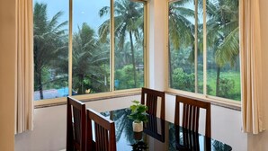 A dining area with a table and chairs, large windows with curtains, and a view of palm trees. - Zuna Stayz Morjim Goa (Morjim)