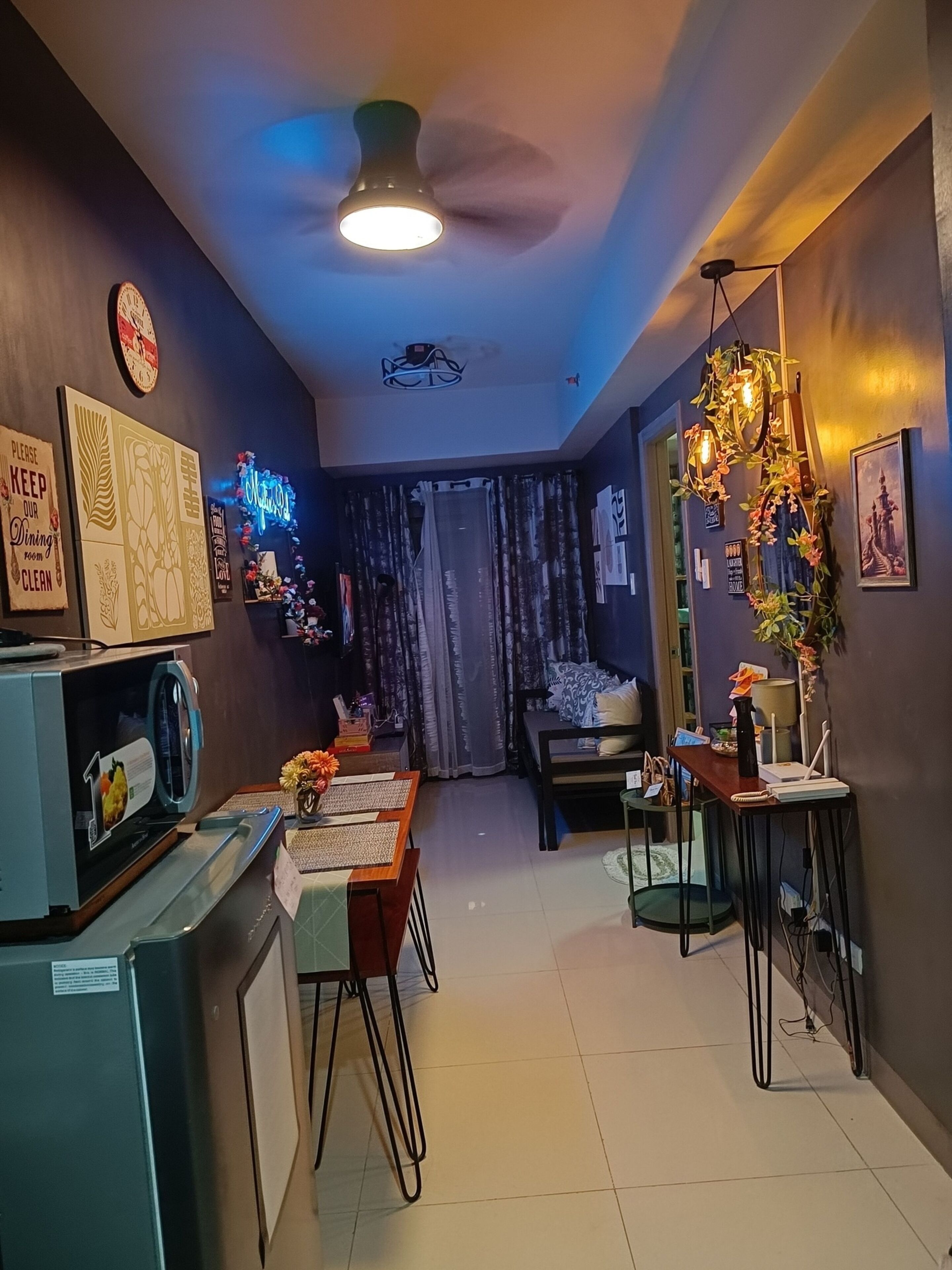 A modern kitchen with a microwave, dining table, and chairs. There is a ceiling fan, a clock, and various wall decorations.