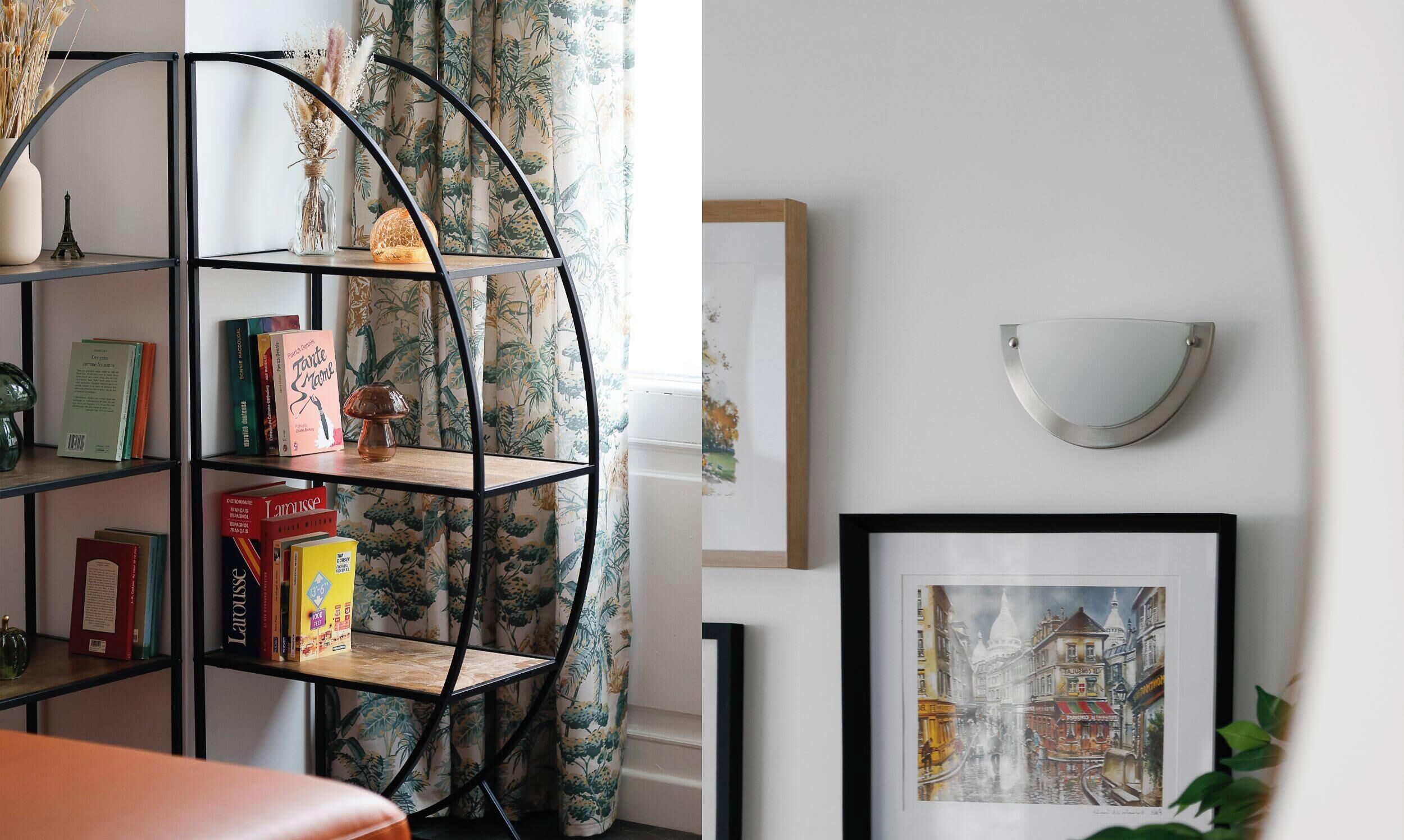 A room with a black metal bookshelf, a framed cityscape painting, and a white wall-mounted light fixture.
