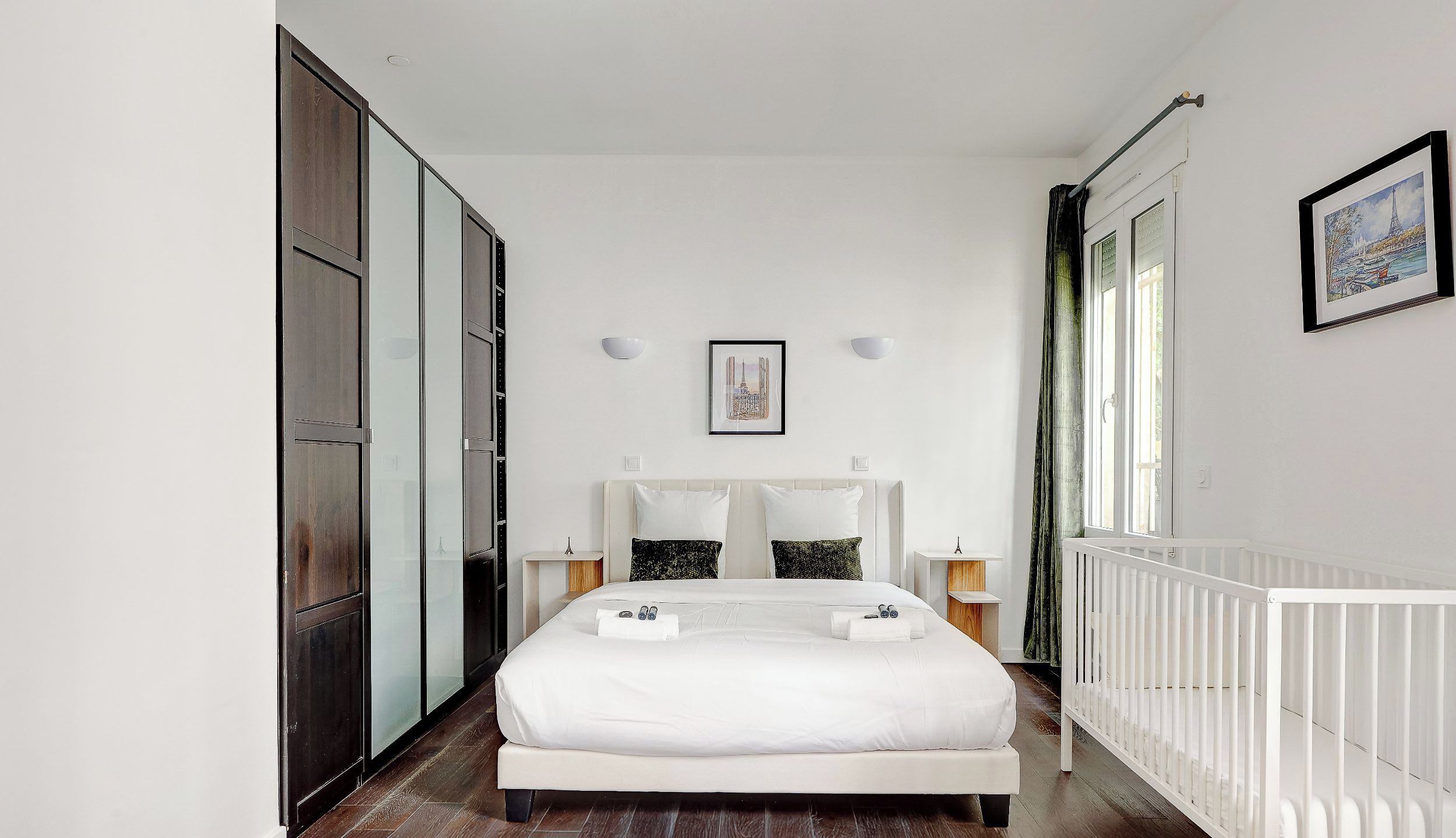 A modern bedroom with a large bed, a white crib, a wardrobe, and a framed picture on the wall.