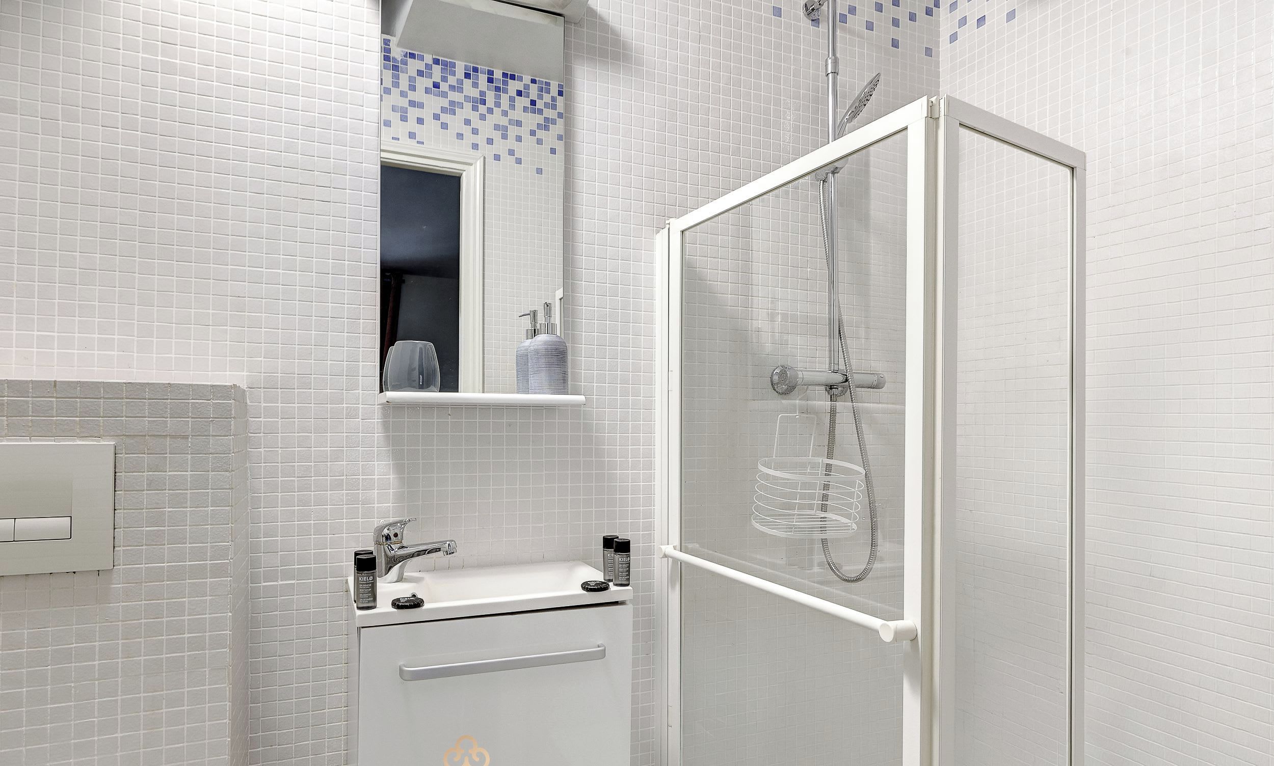 A modern bathroom with white tiled walls, a glass-enclosed shower, a white sink with a mirror above it, and a small shelf with toiletries.