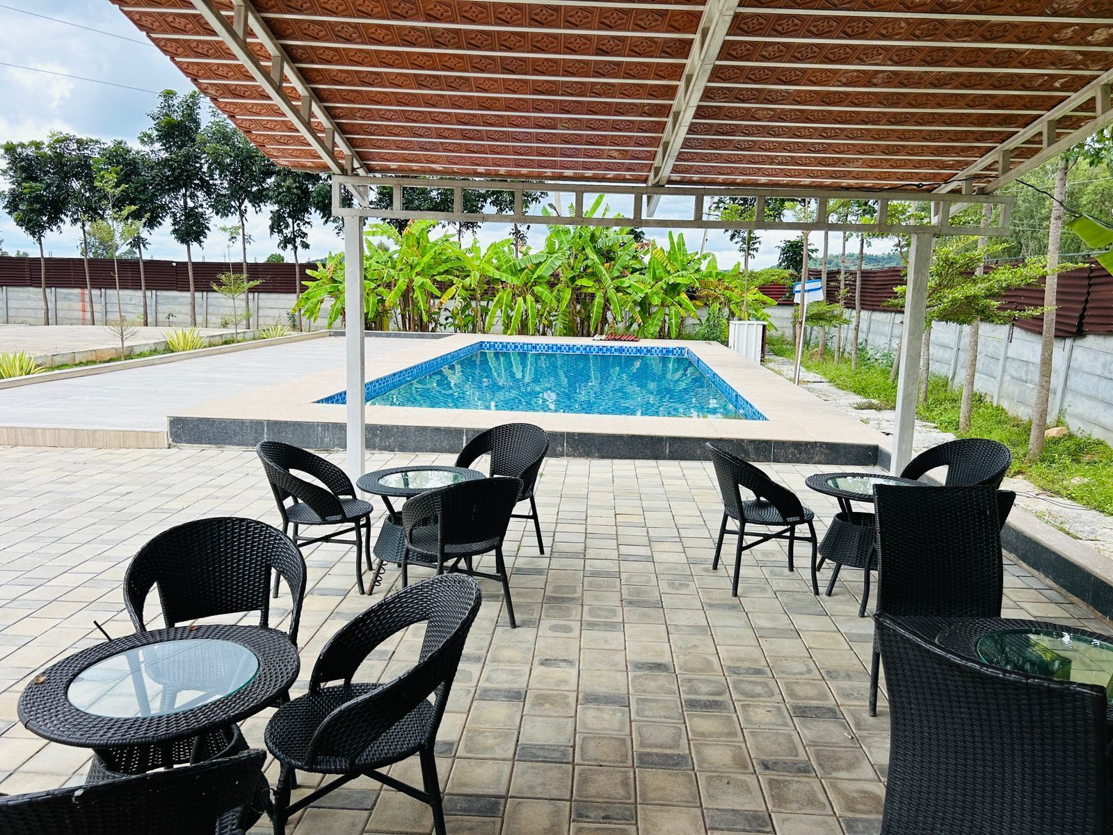Un espacio al aire libre cubierto con piscina, rodeado de sillas y mesas de mimbre negro y abundante vegetación de fondo.